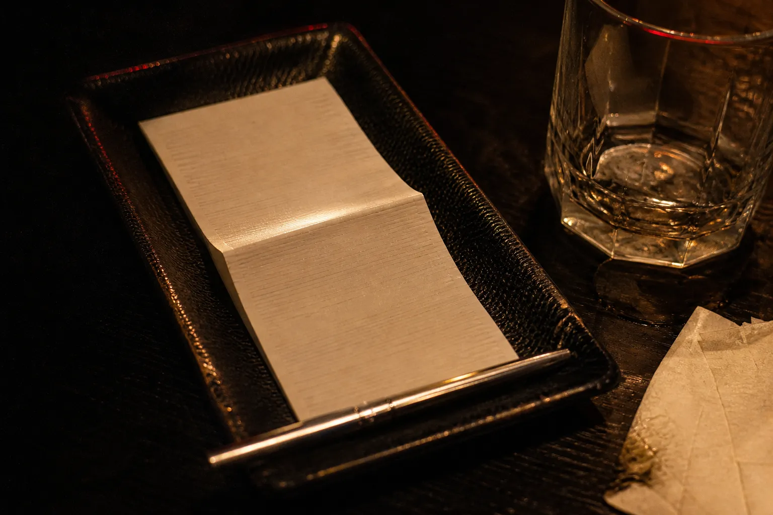 An izakaya bill folded on a black tray beside a glass on a dark wooden table
